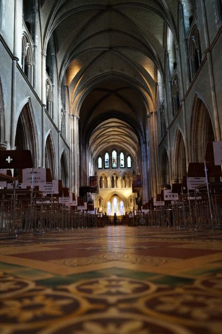 Inside the grand nave of Guildford Cathedral, showing tall stone arches and vaulted ceilings with soft lighting. The floor features patterned tiles, leading towards the altar area illuminated by natur