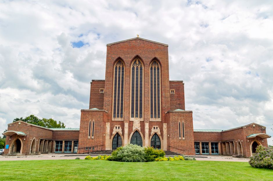 Inside the grand nave of Guildford Cathedral, showing tall stone arches and vaulted ceilings with soft lighting. The floor features patterned tiles, leading towards the altar area illuminated by natur